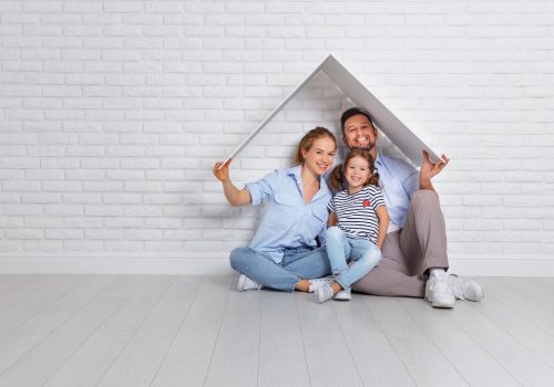 A woman, a man and a child sit on the floor smiling and holding roof shape over their heads